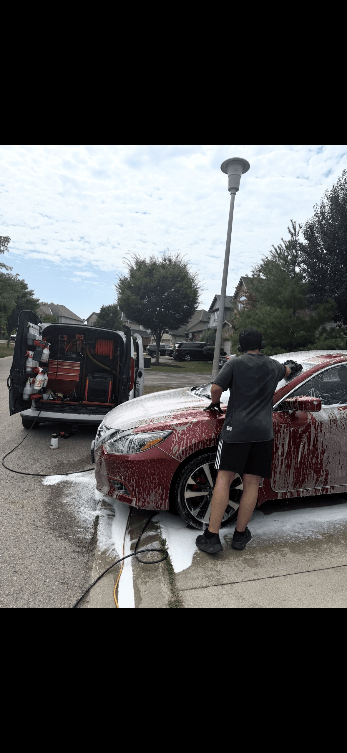 Man washes a soapy red car beside a mobile detailing van on a residential street.