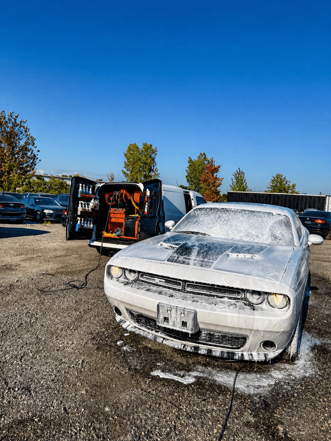 White Dodge Challenger covered in soap foam next to a mobile detailing van outdoors.