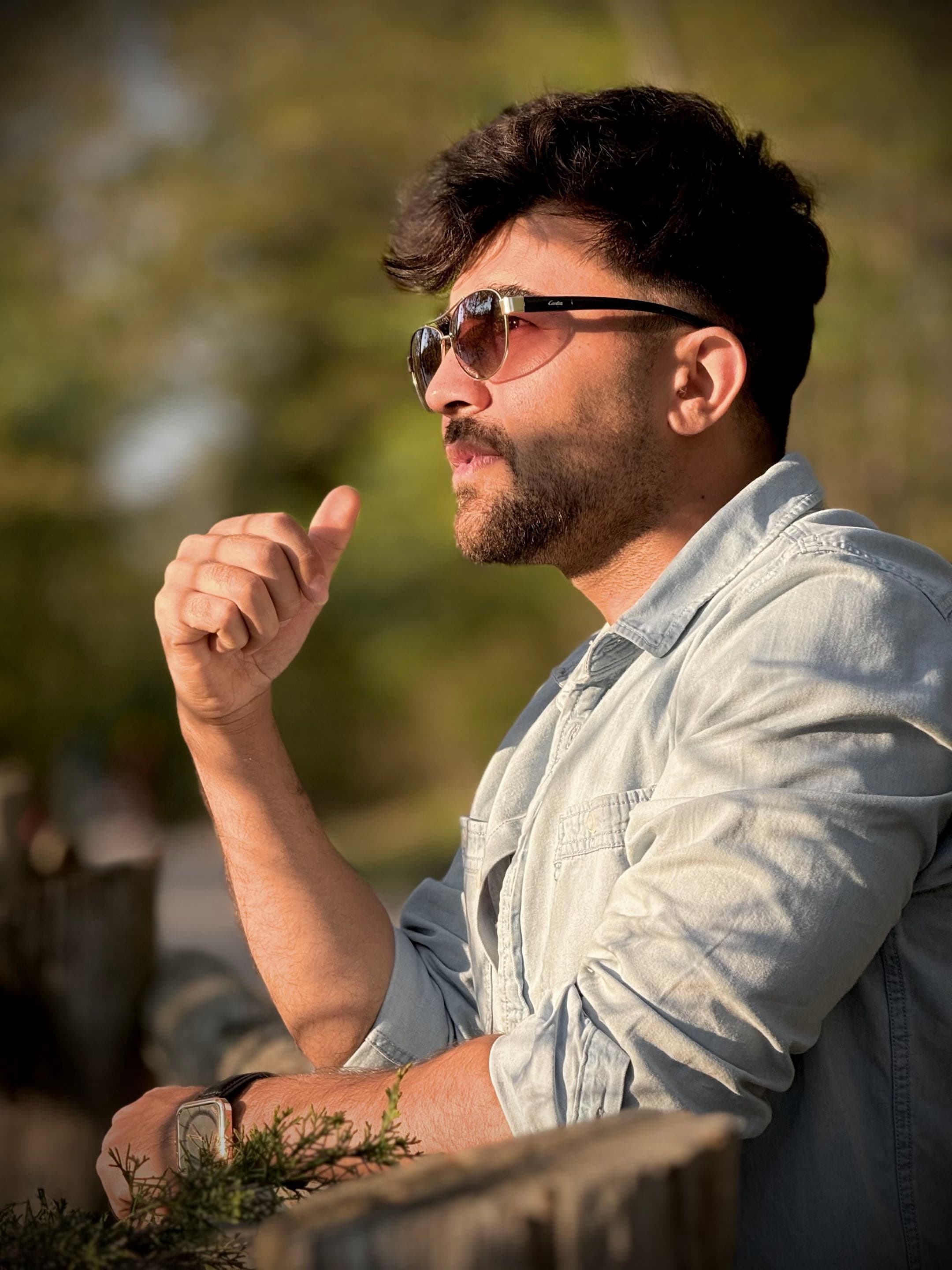 Bearded man in sunglasses and denim shirt looking away, leaning on a fence at sunset.