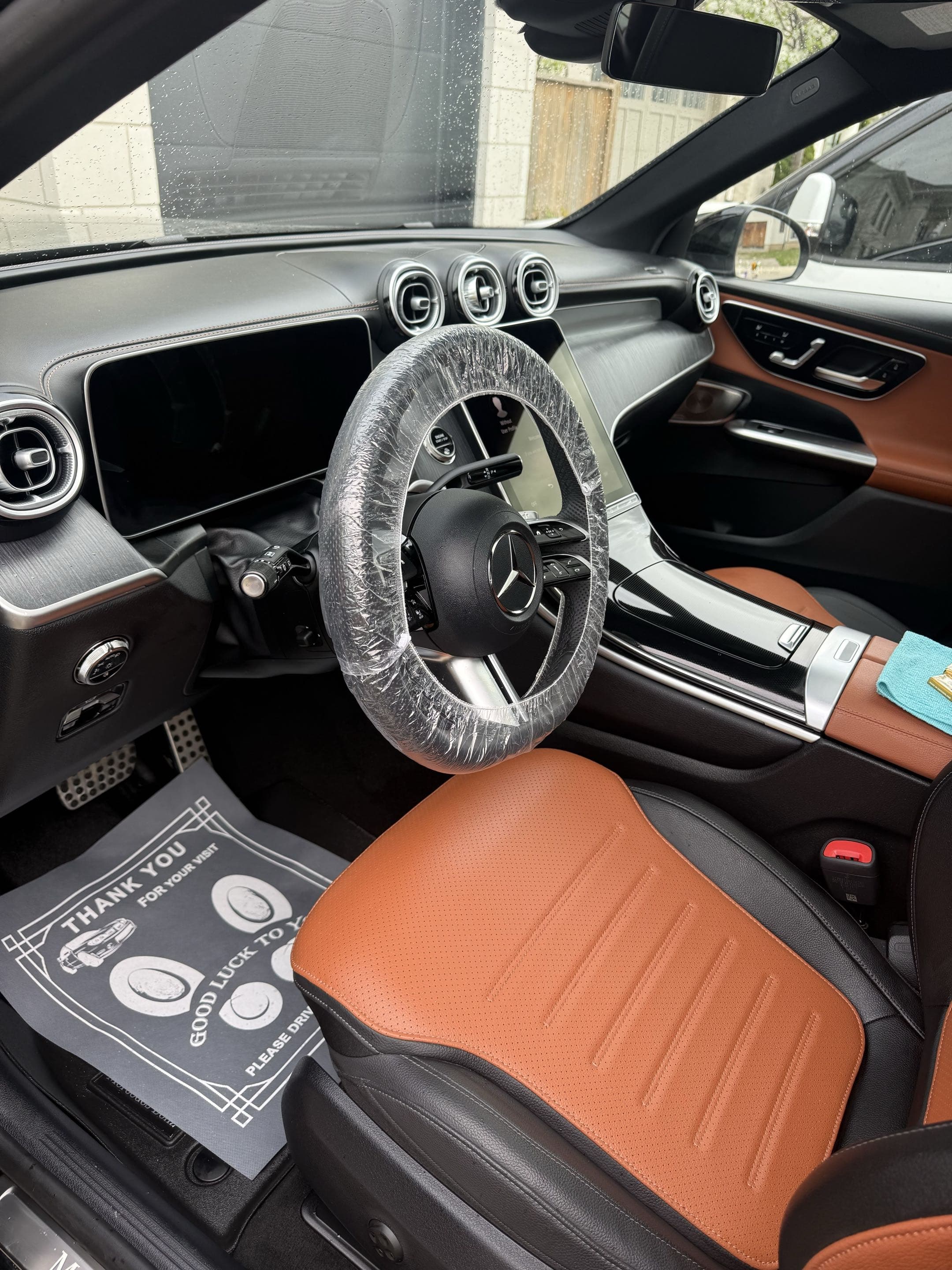 Mercedes-Benz interior featuring a plastic-wrapped steering wheel, digital dashboard, and tan leather seats.