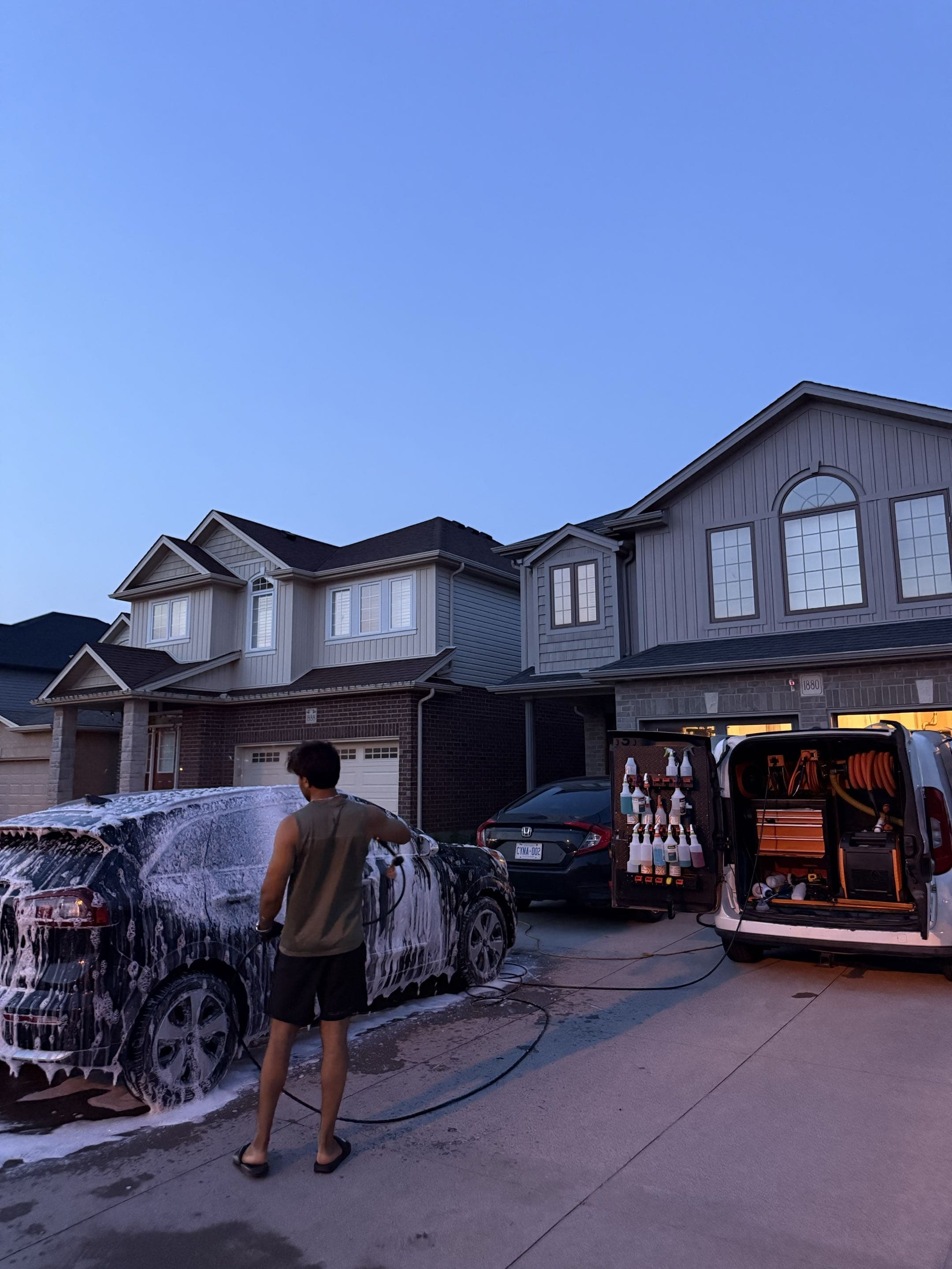 Man washing a foam-covered car in a driveway beside a mobile detailing van at dusk.