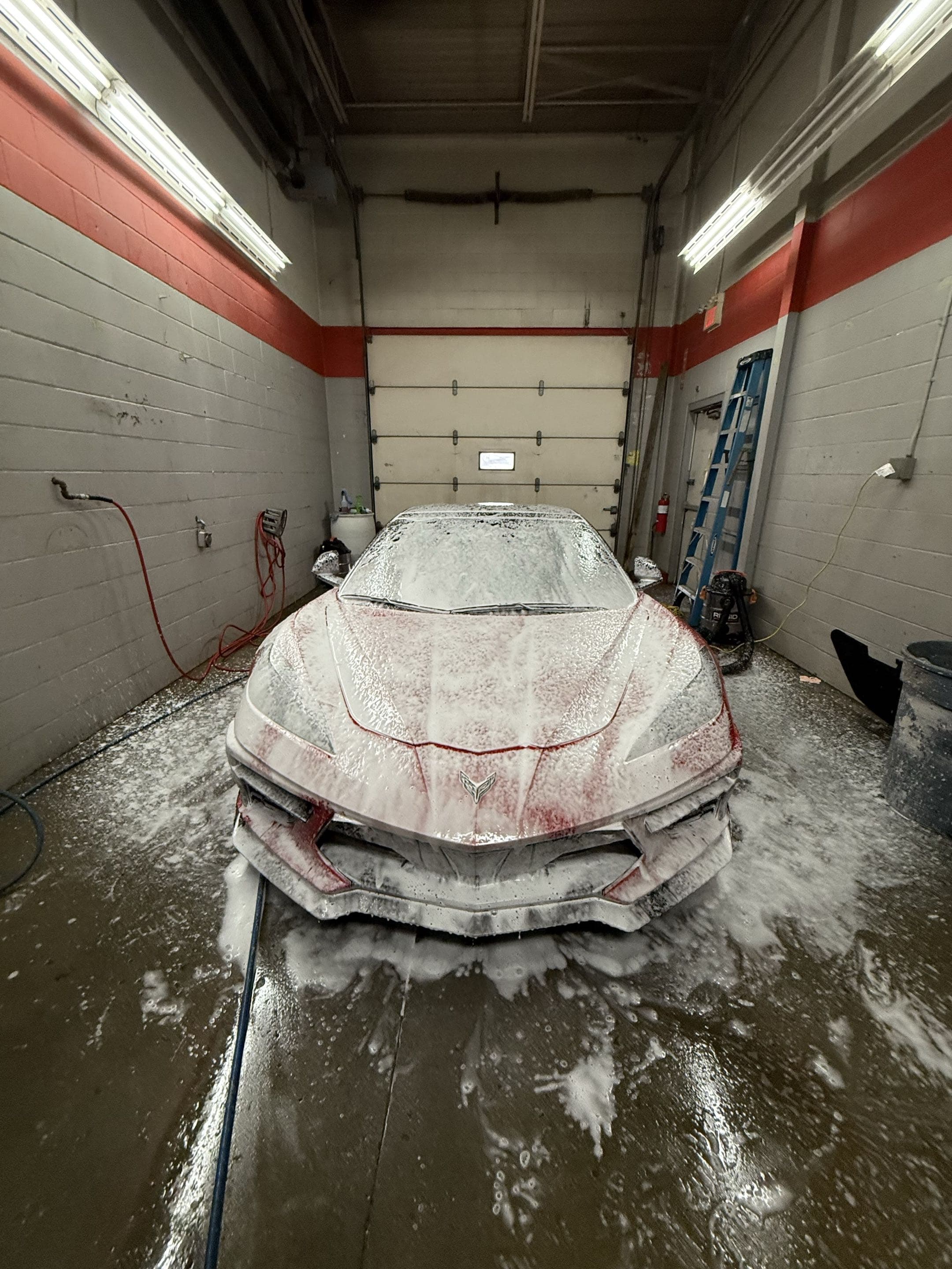 A red Corvette sports car covered in thick white soap foam inside a garage bay.