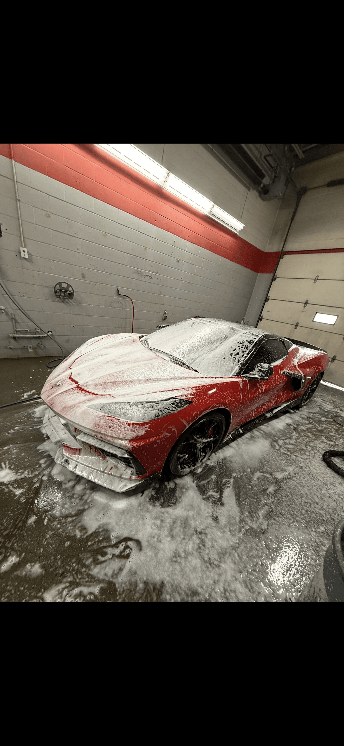 Red sports car covered in thick white soap foam inside a professional car wash bay.